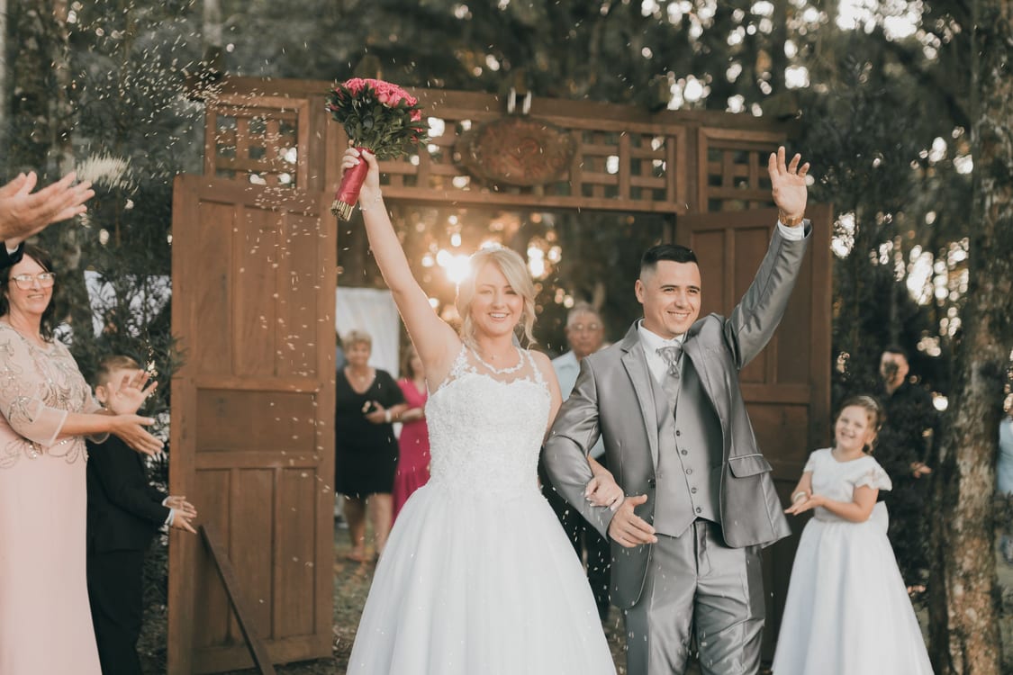 Novios saliendo de la ceremonia al aire libre con los brazos en alto, ella con el ramo, entre invitados que les lanzan arroz