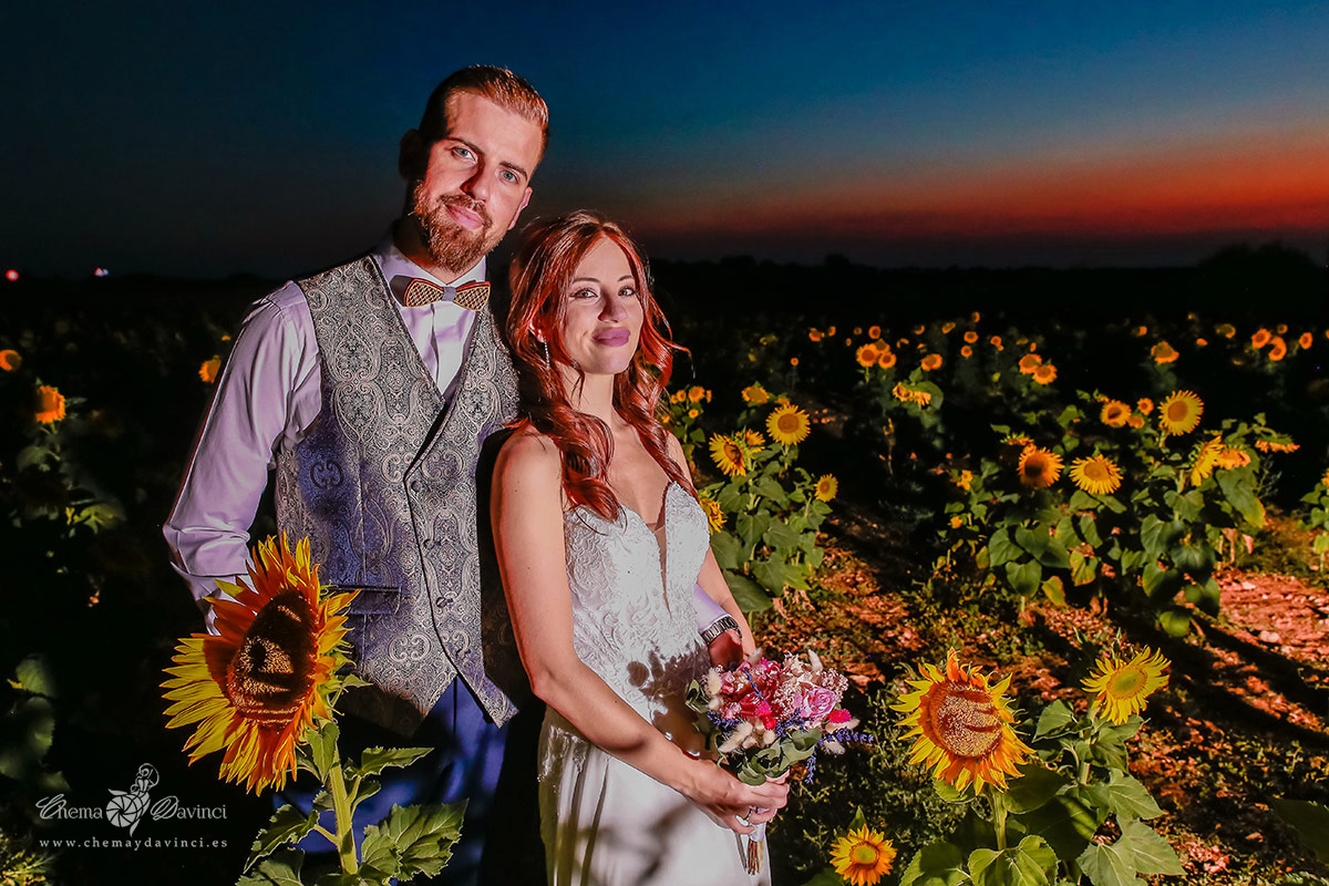 Pareja de recién casados en una sesión postboda en un campo de girasoles al anochecer, ella con el ramo de novia