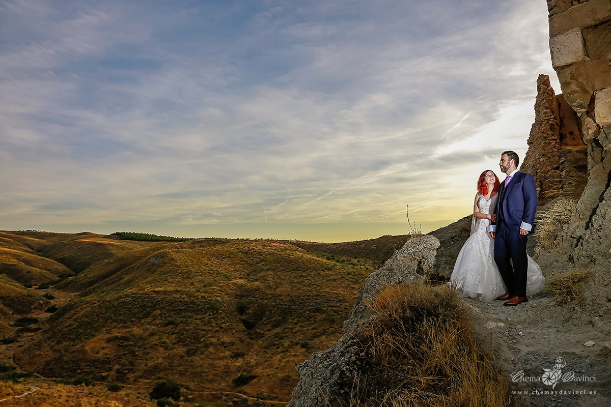 Novios abrazados en lo alto de un cerro rocoso con un valle de colinas al fondo