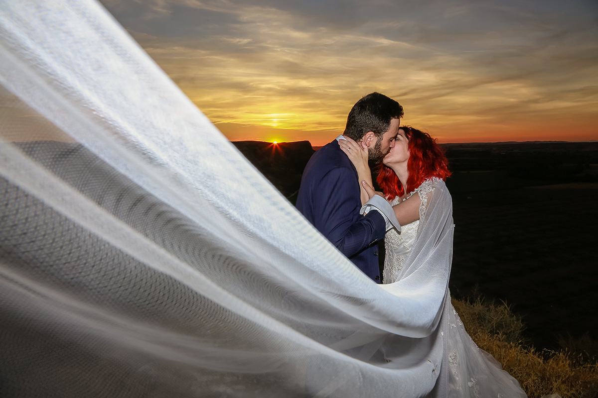 Velo de novia ondeando al viento mientras la pareja se besa al atardecer
